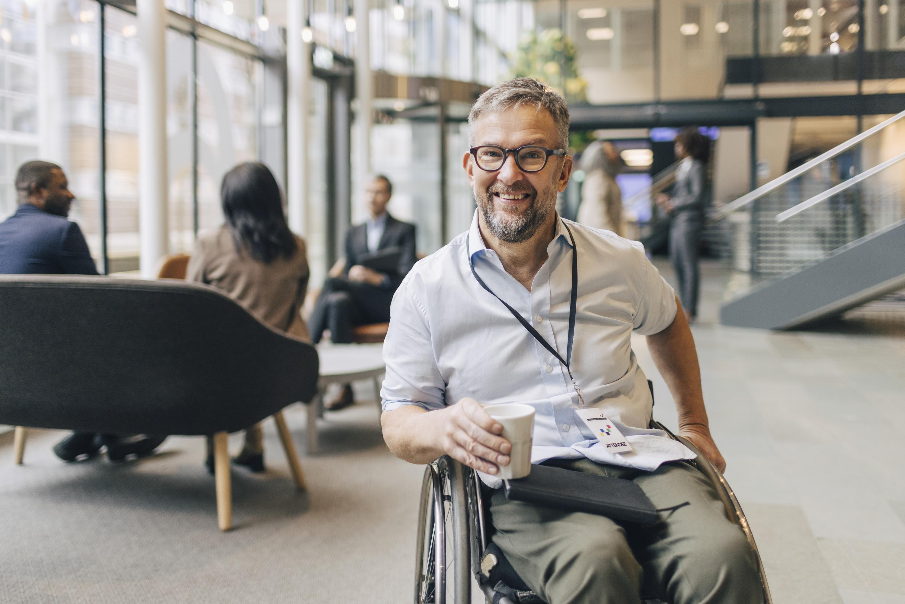 A smiling man in a wheelchair holds a paper cup and wears a name badge in an office.