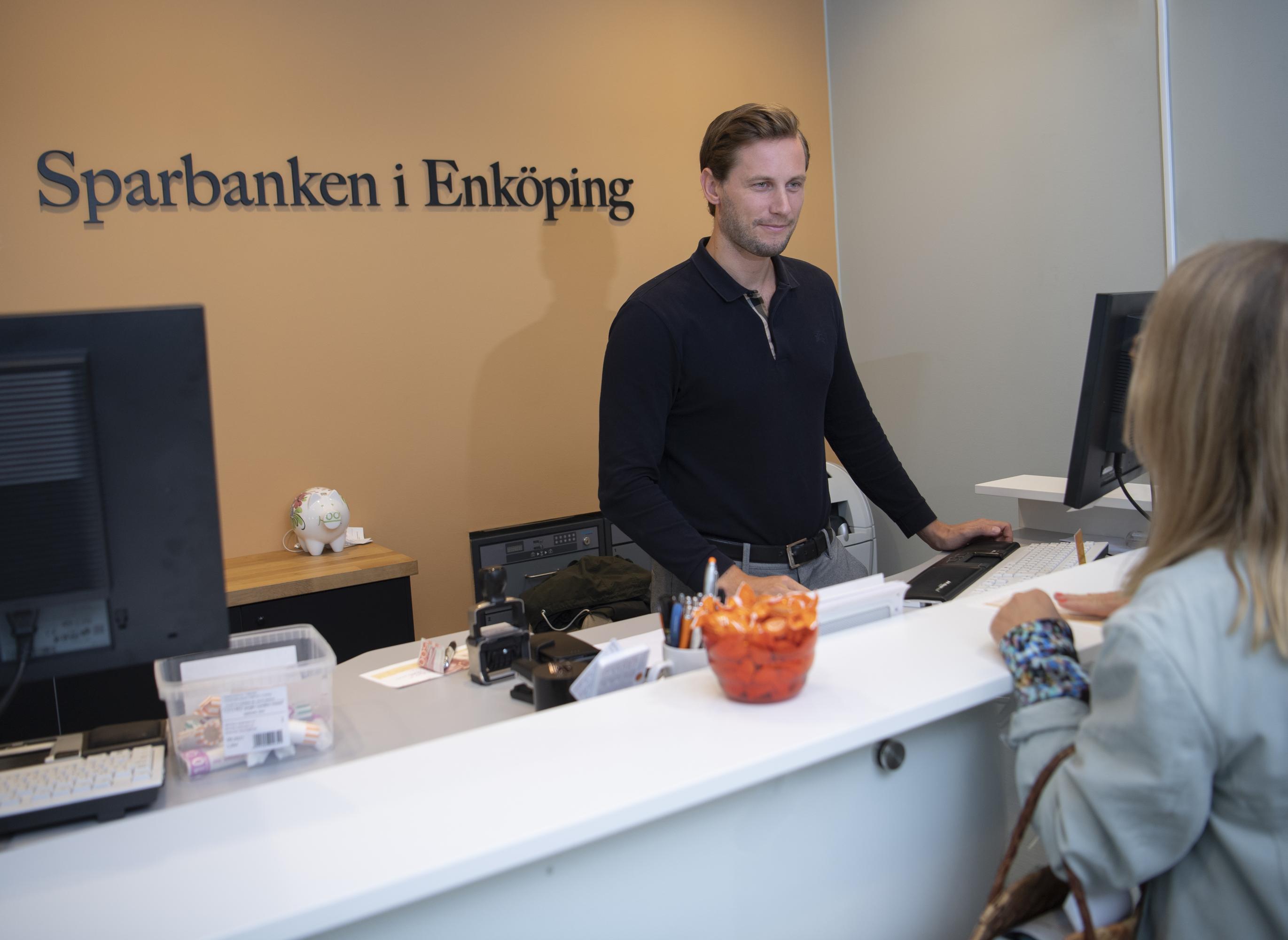 a man in a black shirt is standing next to a computer looking at a customer on the other side of the counter. Sparbanken i Enköping is written on the wall behind him.