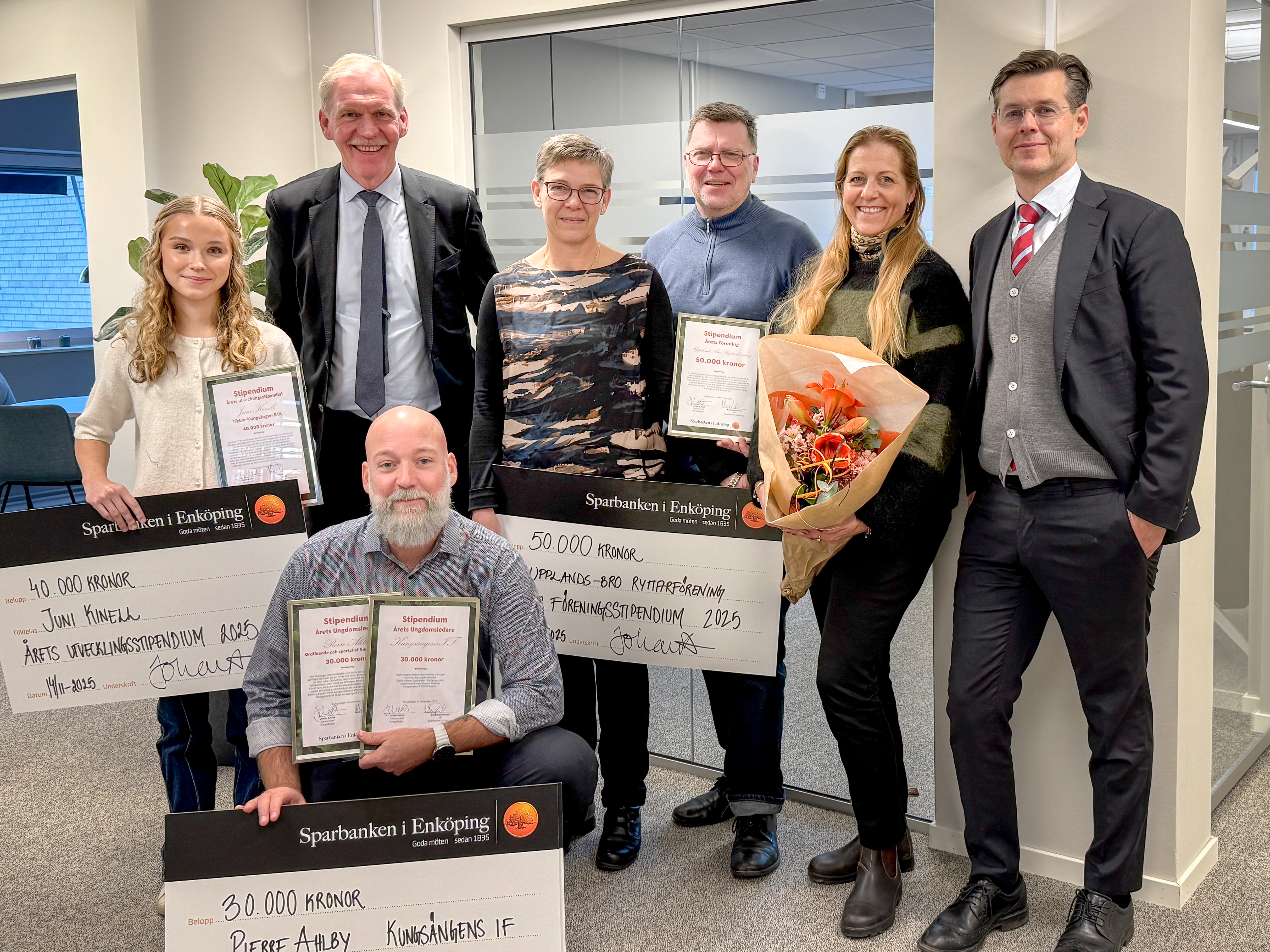 7 personer ler mot kameran, en håller en blombukett tre håller stora checkar från Sparbanken