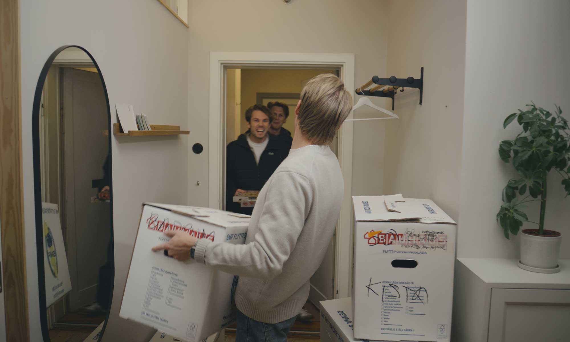 en man håller i en flyttkartong, två unga män på väg in igenom den öppna ytterdörren
